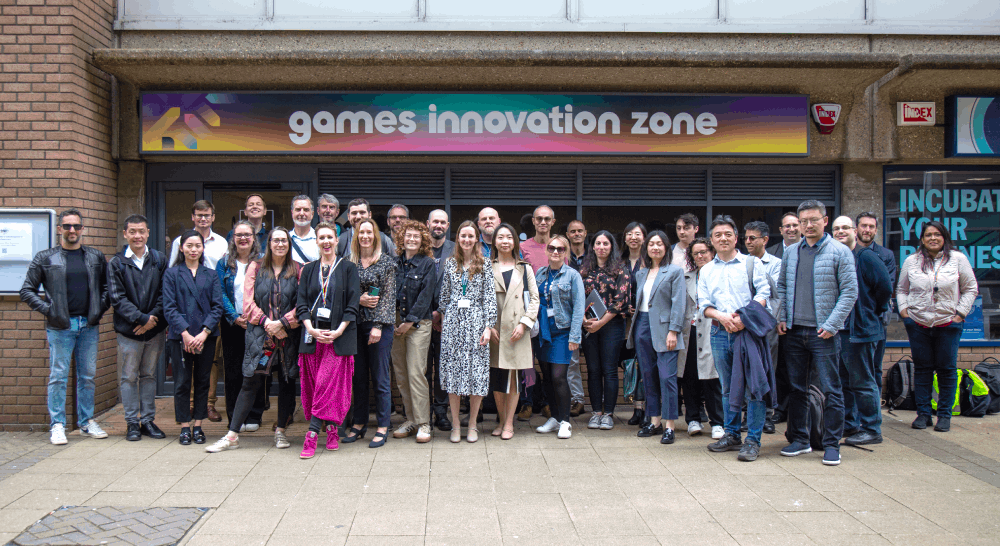 A group of around 30 people stand posing outside of shop front below a brightly coloured board that reads Games Innovation Zone.