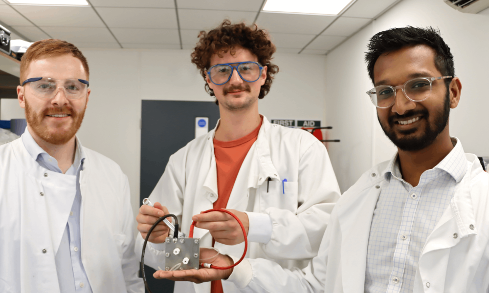 Three men in white lab coats the middle one holds a component with a red and black wire.